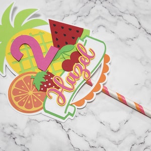 K&ouml;nnte beinhalten: Ein bunter Tortendekor mit Ananas, Wassermelone, Orange, Erdbeere und Kirsche. Der Dekor enth&auml;lt au&szlig;erdem eine rosa Zahl 2 und ein Banner mit der Aufschrift "Hazel". Der Dekor ist an einem rosa-wei&szlig; gestreiften Strohhalm befestigt.