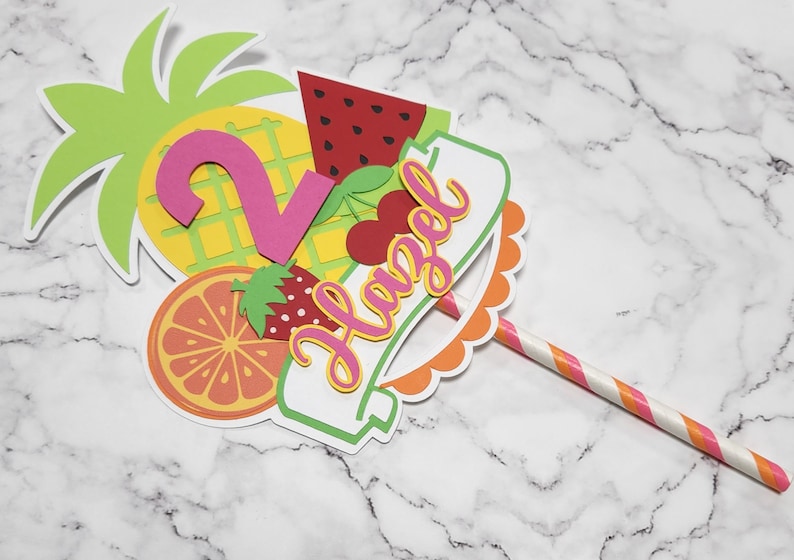 K&ouml;nnte beinhalten: Ein bunter Tortendekor mit Ananas, Wassermelone, Orange, Erdbeere und Kirsche. Der Dekor enth&auml;lt au&szlig;erdem eine rosa Zahl 2 und ein Banner mit der Aufschrift "Hazel". Der Dekor ist an einem rosa-wei&szlig; gestreiften Strohhalm befestigt.