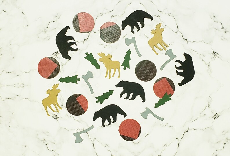 May include: A collection of woodland-themed paper cutouts on a white marble background. The cutouts include bears, moose, pine trees, and axes. The cutouts are in shades of black, brown, green, and yellow.