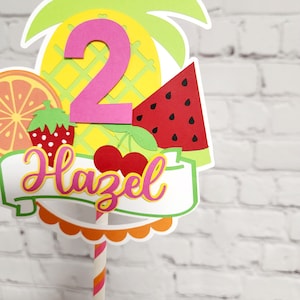K&ouml;nnte beinhalten: Ein bunter, fruchtig gestalteter Tortendekor mit Ananas, Wassermelone, Orange, Erdbeere und Kirschen. Die Zahl "2" ist in Pink und der Name "Hazel" ist in Gelb mit einem wei&szlig;en Banner. Der Dekor ist auf einem gestreiften Strohhalm.
