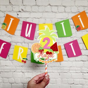 K&ouml;nnte beinhalten: Ein bunter, fruchtiger Tortendekor mit Ananas, Wassermelone, Orange und Erdbeeren. Der Dekor hat eine rosa Zahl "2" und den Namen "Hazel" in rosa Schrift. Der Dekor ist an einem gestreiften Strohhalm befestigt.