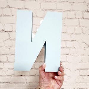 以下が含まれることがあります： 白レンガの壁を背景に、水色の立体的な文字「N」が手で持たれています。文字はしっかりとした素材でできており、マットな仕上がりです。文字を持っている人は、ベージュのバンドの時計をしています。