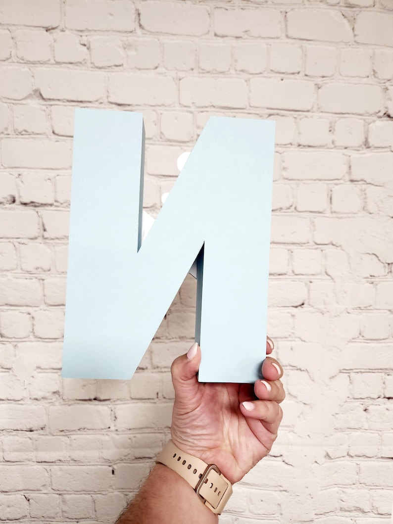 以下が含まれることがあります： 白レンガの壁を背景に、水色の立体的な文字「N」が手で持たれています。文字はしっかりとした素材でできており、マットな仕上がりです。文字を持っている人は、ベージュのバンドの時計をしています。