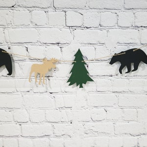 May include: A woodland-themed banner featuring cut-outs of a moose, a pine tree, and two bears. The banner is strung with twine and is hanging on a white brick wall.