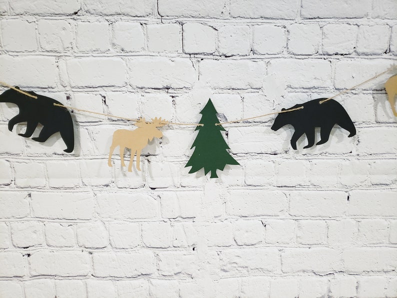 May include: A woodland-themed banner featuring cut-outs of a moose, a pine tree, and two bears. The banner is strung with twine and is hanging on a white brick wall.