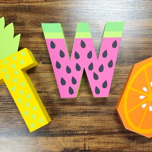 May include: Three wooden letters spelling out the word "TWO" decorated with fruit designs. The letter "T" is yellow and green and resembles a pineapple. The letter "W" is pink and green and resembles a watermelon. The letter "O" is orange and white and resembles an orange slice.