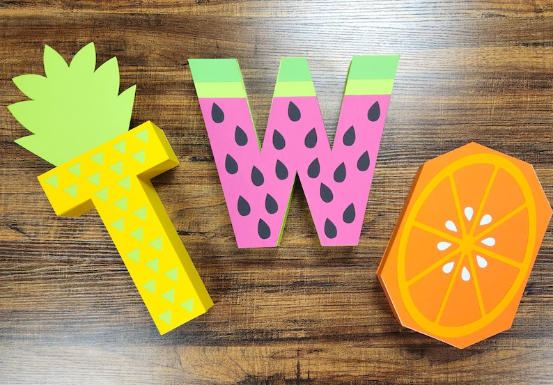 May include: Three wooden letters spelling out the word "TWO" decorated with fruit designs. The letter "T" is yellow and green and resembles a pineapple. The letter "W" is pink and green and resembles a watermelon. The letter "O" is orange and white and resembles an orange slice.