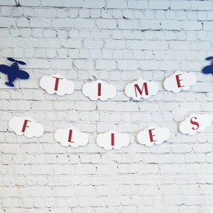 May include: White cloud-shaped paper cutouts with red letters spelling "TIME FLIES" are strung together with blue airplane cutouts on a white brick wall background.