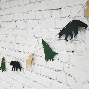 May include: A woodland-themed paper banner with alternating green trees, brown moose, and black bears. The banner is strung on a white brick wall.