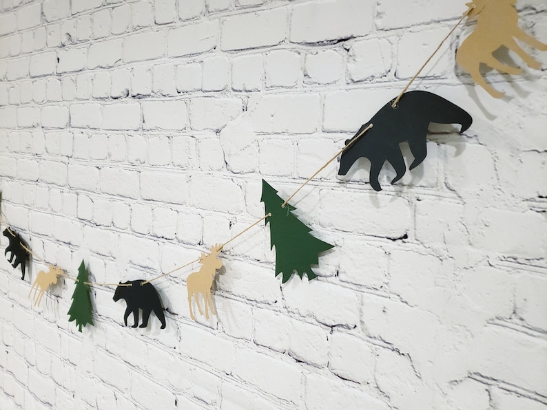 May include: A woodland-themed paper banner with alternating green trees, brown moose, and black bears. The banner is strung on a white brick wall.