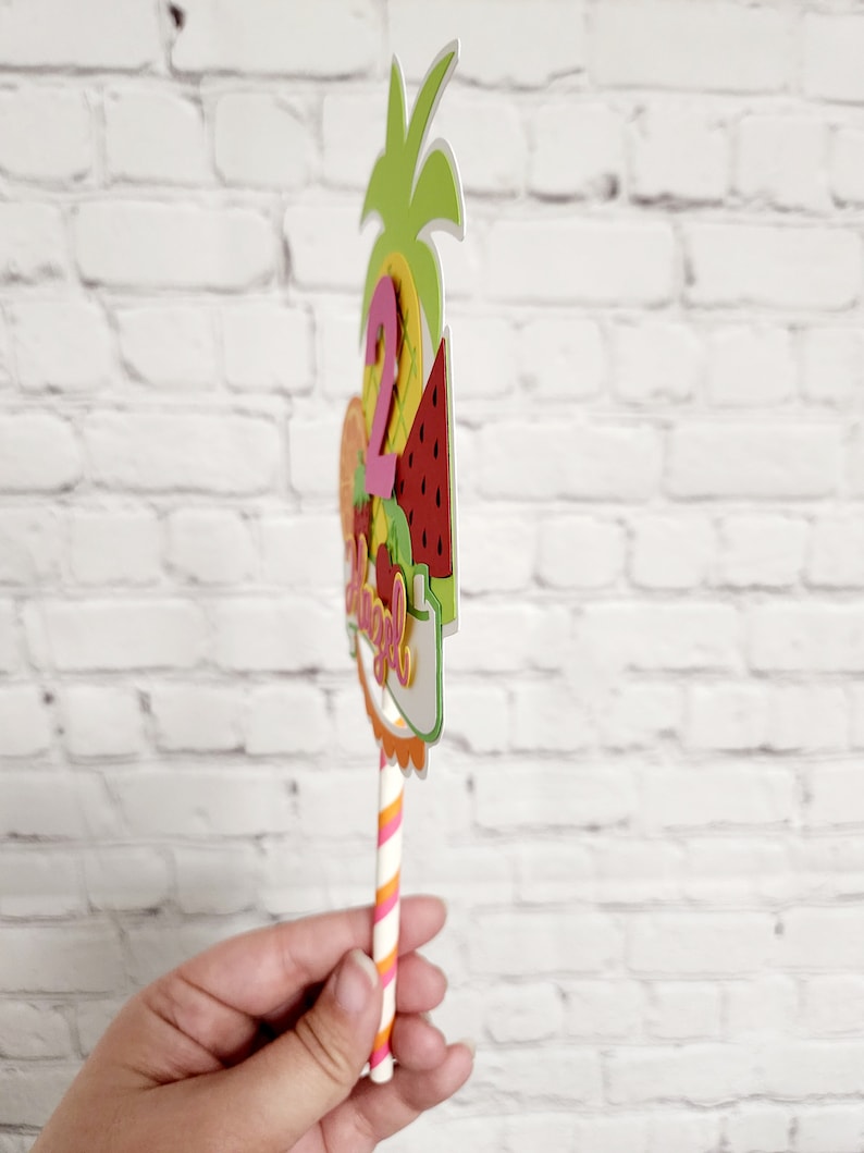 K&ouml;nnte beinhalten: Ein bunter Obst-Kuchentopper mit Ananas, Wassermelone und der Zahl "2" auf einem gestreiften Strohhalm. Der Topper sagt "Happy 2nd Birthday".