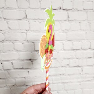 K&ouml;nnte beinhalten: Ein bunter Tortendekor mit Ananas-, Wassermelonen-, Orangen- und Erdbeerdesign. Der Dekor zeigt die Zahl "2" und das Wort "Happy" in einer rosa und orangen Schriftart. Der Dekor ist an einem gestreiften Strohhalm befestigt.