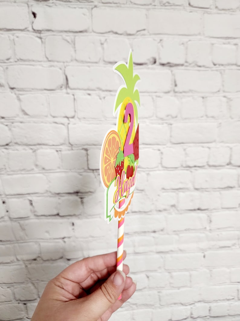 K&ouml;nnte beinhalten: Ein bunter Tortendekor mit Ananas-, Wassermelonen-, Orangen- und Erdbeerdesign. Der Dekor zeigt die Zahl "2" und das Wort "Happy" in einer rosa und orangen Schriftart. Der Dekor ist an einem gestreiften Strohhalm befestigt.