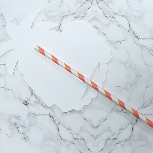 K&ouml;nnte beinhalten: Ein wei&szlig;er Papierschnitt eines Palmenbaums mit einem rosa-orangefarbenen, gestreiften Papierstrohhalm, der durch die Mitte gesteckt ist.