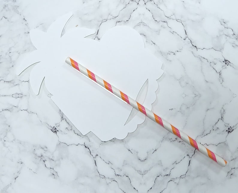 K&ouml;nnte beinhalten: Ein wei&szlig;er Papierschnitt eines Palmenbaums mit einem rosa-orangefarbenen, gestreiften Papierstrohhalm, der durch die Mitte gesteckt ist.