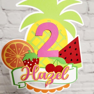 K&ouml;nnte beinhalten: Ein bunter Tortendekor mit Ananas, Wassermelone, Orange, Erdbeere und Kirschen. Die Zahl 2 ist in Pink und der Name Hazel ist in Pink und Gelb auf einem wei&szlig;en Banner. Der Topper ist auf einem gestreiften Strohhalm.