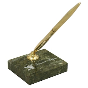 May include: A gold-colored pen set on a green marble base. The base has the text "Splatter Media" and a silver ink splatter design. The pen is angled upwards, ready for use.