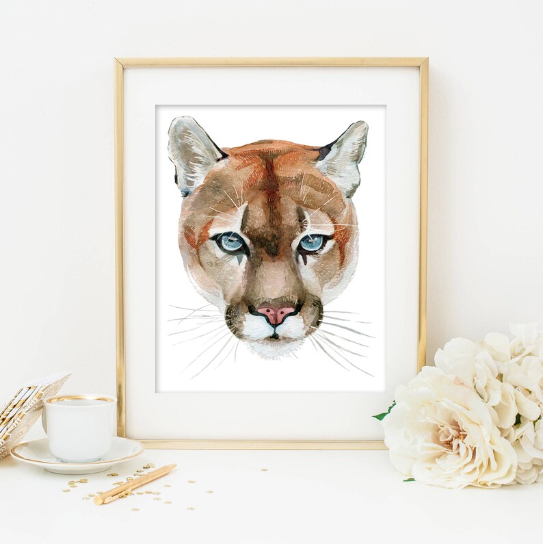 May include: A watercolor painting of a mountain lion's face. The lion has brown fur, blue eyes, and a pink nose. The painting is framed in a gold frame.