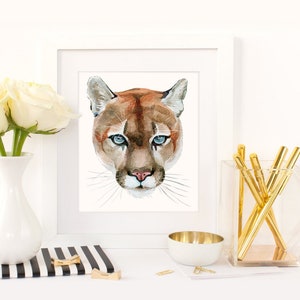 May include: Watercolor painting of a mountain lion's face with blue eyes. The painting is framed in a white frame and is sitting on a table with a white vase of white roses and a gold pen holder.