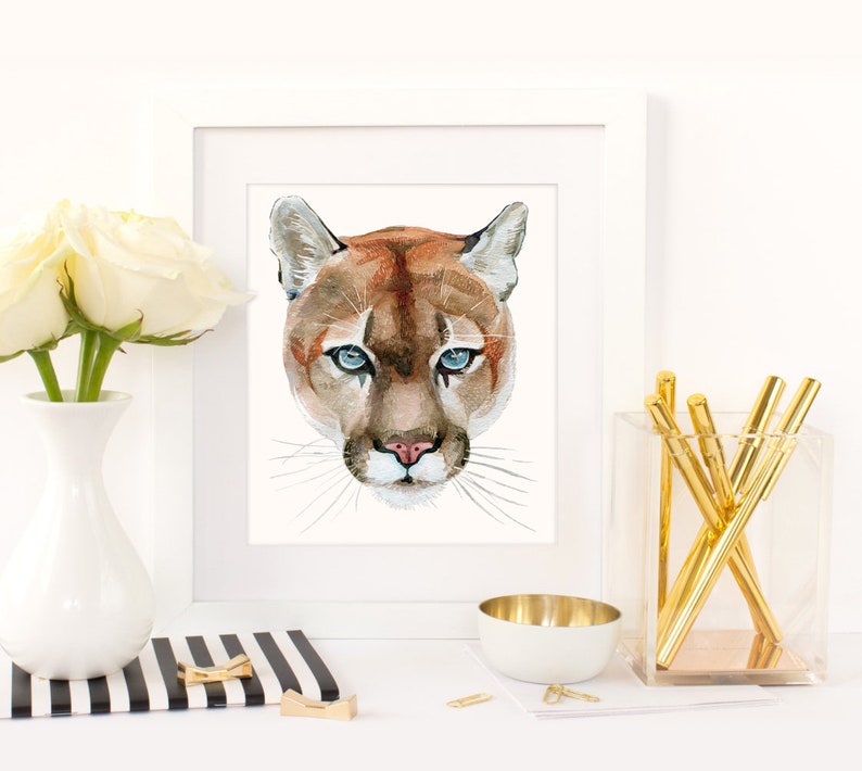 May include: Watercolor painting of a mountain lion's face with blue eyes. The painting is framed in a white frame and is sitting on a table with a white vase of white roses and a gold pen holder.