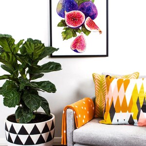 May include: A framed watercolor painting of purple and pink figs with green leaves. The painting is hanging on a white wall above a gray armchair with orange and white patterned pillows.