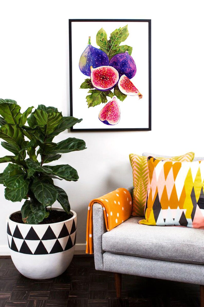 May include: A framed watercolor painting of purple and pink figs with green leaves. The painting is hanging on a white wall above a gray armchair with orange and white patterned pillows.