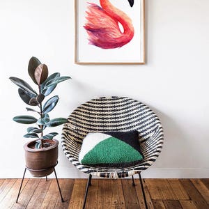 May include: A framed watercolor painting of a pink flamingo with a white background. The painting is hanging on a white wall above a black and white striped wicker chair with a green and white pillow.