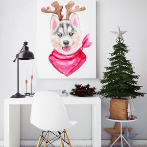 May include: A watercolor painting of a husky puppy wearing a red bandana and reindeer antlers. The painting is framed and hanging on a white wall. A small Christmas tree is in the background.