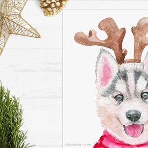 May include: A watercolor painting of a husky puppy wearing a red and white reindeer costume with antlers. The puppy is looking at the viewer with its tongue sticking out.