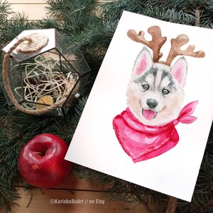 May include: Watercolor painting of a husky puppy wearing a pink bandana and reindeer antlers. The puppy is looking at the viewer with a happy expression.