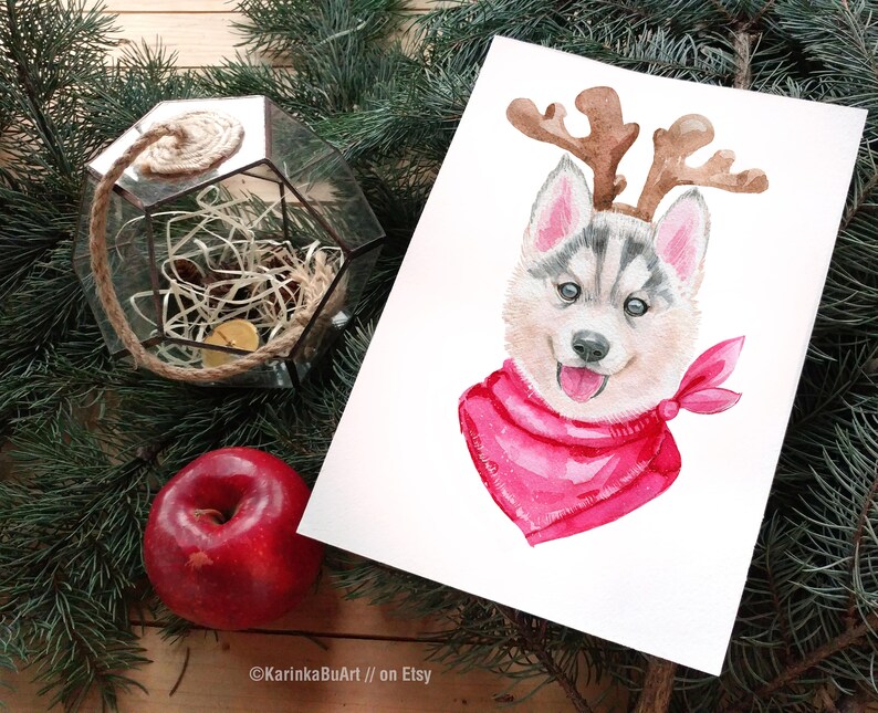 May include: Watercolor painting of a husky puppy wearing a pink bandana and reindeer antlers. The puppy is looking at the viewer with a happy expression.