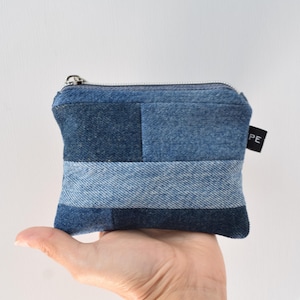 May include: A small rectangular pouch constructed from patchwork denim fabric in varying shades of blue. The pouch features a horizontal zipper closure along the top edge and a small black fabric tag on the side with the partial white text PE.