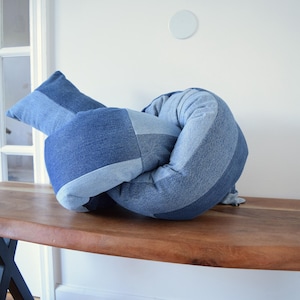 May include: A knot-shaped decorative pillow constructed from various shades of blue denim fabric. The pillow features a patchwork design with alternating light and dark blue denim panels, creating a textured, knotted appearance in a soft, stuffed form.