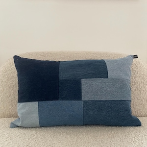 Pode incluir: Uma almofada retangular de ganga azul com um design de patchwork. A almofada é feita de diferentes tons de ganga azul e tem um visual simples e moderno.