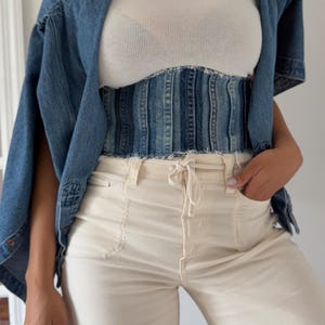 Puede incluir: Un corsé de mezclilla azul con paneles verticales y bordes deshilachados sobre una blusa blanca acanalada y pantalones color crema con cordón, junto con una camisa de mezclilla azul sobre los hombros para completar el conjunto.
