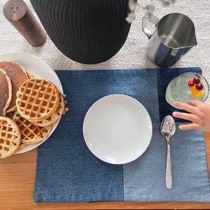May include: A blue denim placemat with a white plate, a silver spoon, and a glass of water with cranberries and an orange slice. The placemat is on a wooden table with a plate of waffles and a black vase with white flowers.