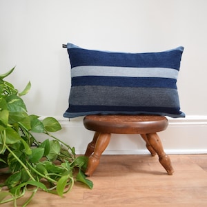 May include: A rectangular decorative pillow with horizontal stripes in shades of blue and gray. The pillow is placed on a small, round, wooden stool with three legs. Green leafy vines are in the foreground.