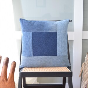 May include: A square denim pillow with a patchwork design in various shades of blue. The pillow is placed on a black chair with a woven seat. The pillow's design features a darker blue square in the center, surrounded by lighter blue denim.