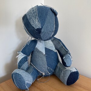 Könnte beinhalten: Ein handgefertigter Teddybär aus Patchwork-Denim. Der Bär ist hauptsächlich blau mit verschiedenen Denim-Farbtönen. Er hat einen runden Kopf, kleine Ohren und gestickte Details. Der Bär sitzt auf einer Holzoberfläche.