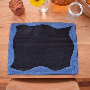 May include: A denim placemat with a dark center and a wavy blue border. The placemat is on a wooden table, with a yellow flower in a glass and other decorative items.