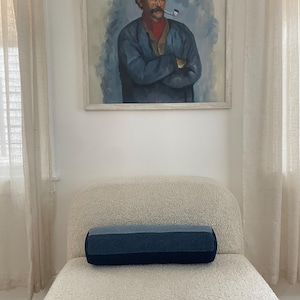 May include: A white, fluffy,  upholstered bench with a blue and black bolster pillow. A framed painting of a man with a pipe hangs on the wall behind the bench.