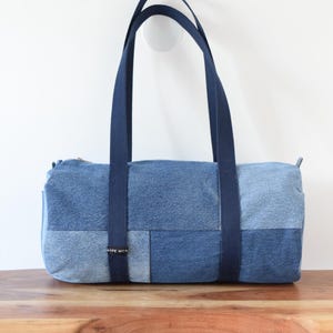 May include: A cylindrical duffel bag constructed from patchwork denim in various shades of blue. The bag features two dark blue fabric handles and a small black label with white text on the front panel. The design includes a top zipper closure.