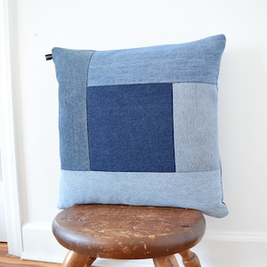 May include: Square denim pillow with a patchwork design in various shades of blue. The pillow is placed on a small, round wooden stool with carved details. The pillow's design features a dark blue square in the center.