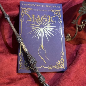 Elegant deep purple book titled The Proficiently Practical Compendium of Magic with gold detailing and radiant hand design. Paired with a detailed dark wand and set on a rich burgundy fabric backdrop. A decorative stand is visible on the right.