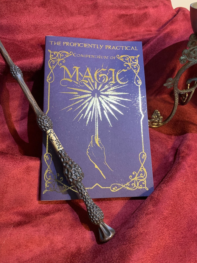 Elegant deep purple book titled The Proficiently Practical Compendium of Magic with gold detailing and radiant hand design. Paired with a detailed dark wand and set on a rich burgundy fabric backdrop. A decorative stand is visible on the right.