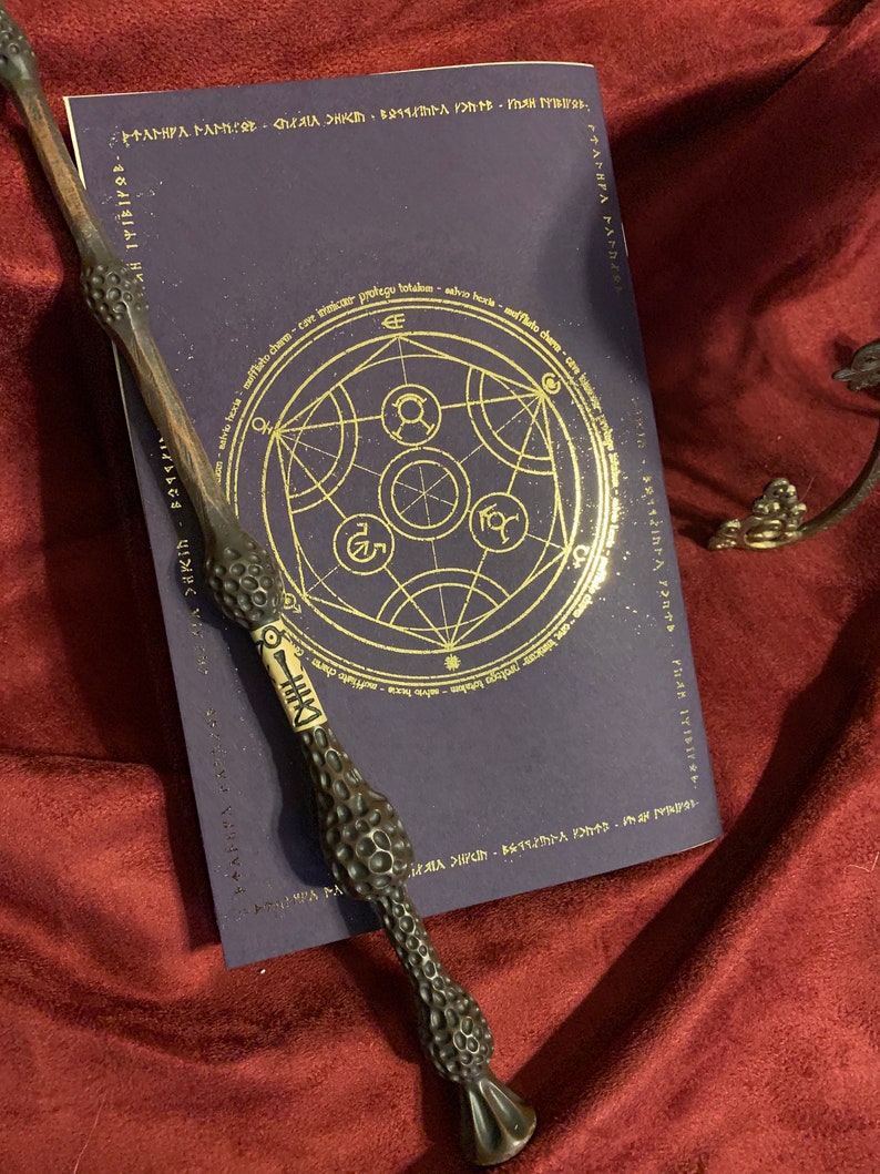 May include: A dark brown, textured wand with a light-colored central band featuring black markings, resting diagonally across a dark purple book. The book cover displays a gold circular geometric design and text including 'salvio hexia'.