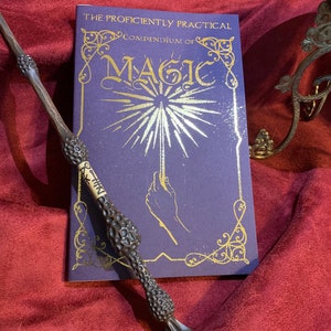 May include: A dark brown, textured wand with a light-colored band featuring black symbols, placed next to a purple book titled The Proficiently Practical Compendium of Magic, which displays gold lettering and a graphic of a hand holding a glowing wand.
