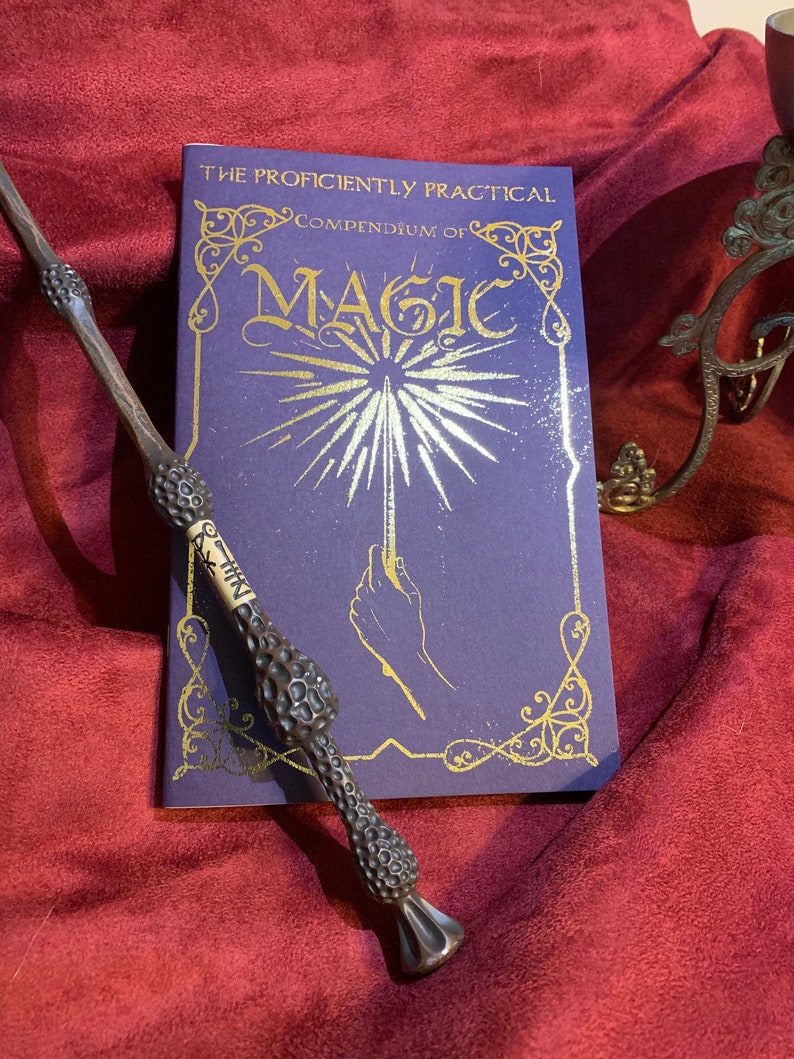 May include: A dark brown, textured wand with a light-colored band featuring black symbols, placed next to a purple book titled The Proficiently Practical Compendium of Magic, which displays gold lettering and a graphic of a hand holding a glowing wand.