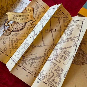 Folded parchment displaying intricate blueprints of a magical castle's interior. Details include rooms, hallways, and notes about secret passages. Centered is a banner that reads Secrets on Every Page. Background features a plush red fabric.