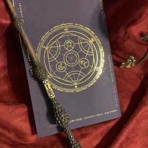 Back cover of a deep purple book, displaying intricate gold celestial and alchemical symbols. Alongside is a detailed dark wand resting on a luxurious burgundy fabric. To the right, a hint of an ornate stand can be seen.